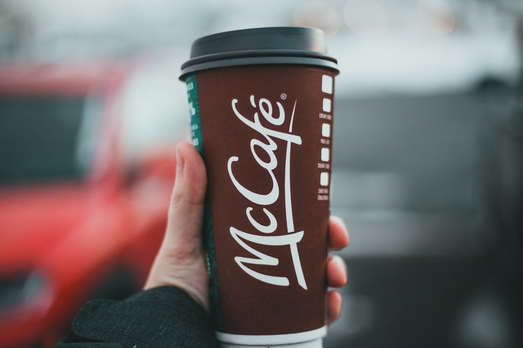 Pessoa segurando copo marrom escrito McCafé. O logotipo da marca registrada é o título junto com um símbolo R